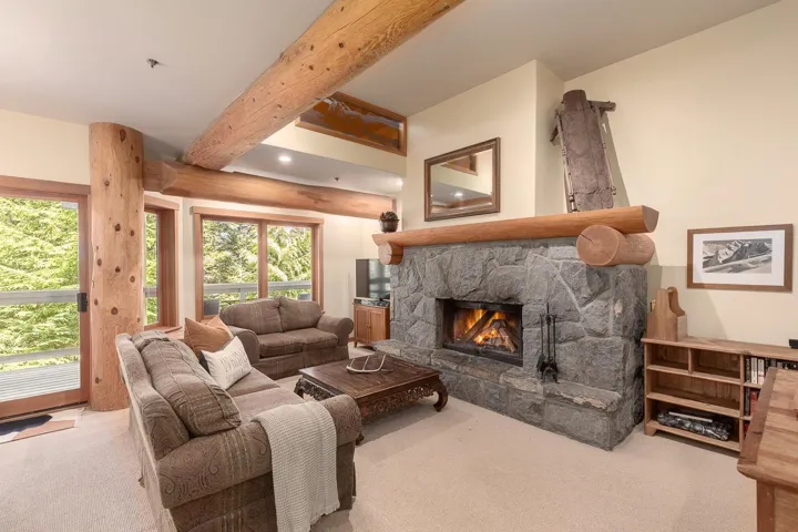 Spacious living area featuring a stone-clad fireplace with a prominent log mantel, exposed wood beams, and large windows