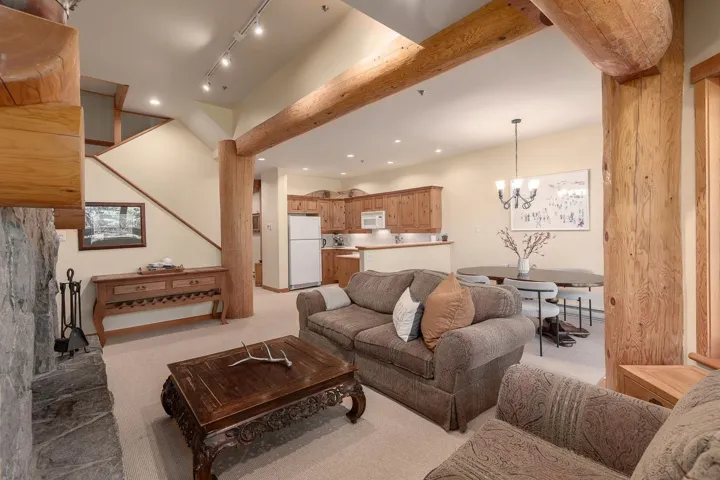 Stone fireplace with log mantel, open-concept living area with exposed wooden beams, staircase with wood railing, recessed lighting, and kitchen with wood cabinetry