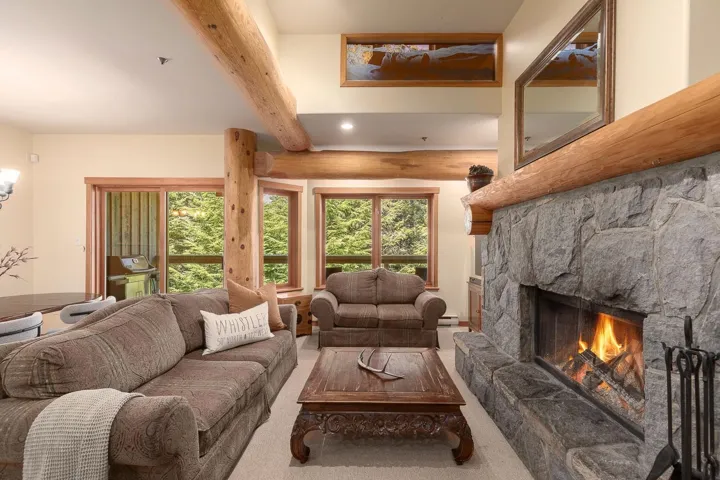 Spacious living area featuring a stone fireplace with a substantial wood mantel, exposed log beams, recessed lighting, and large windows with wood trim