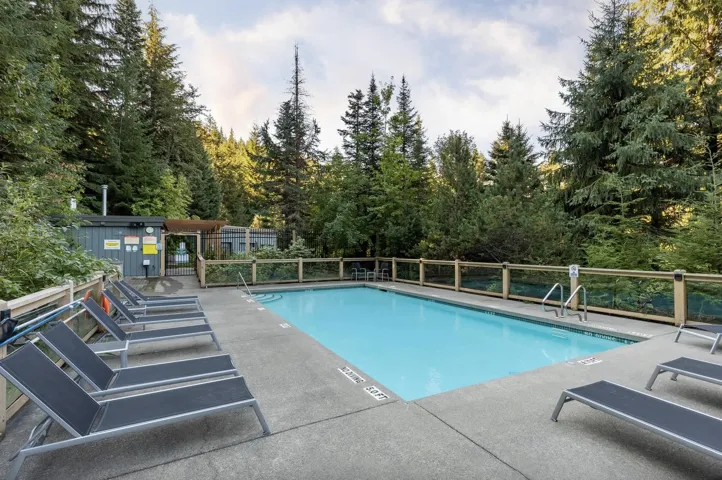 Outdoor swimming pool featuring a concrete deck, a transparent safety fence with wood-finish railing, and perimeter landscaping with mature evergreen trees