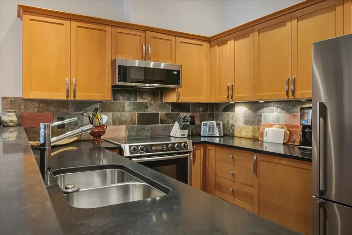 Kitchen featuring decorative backsplash, stainless steel appliances, brown cabinetry, and dark stone counters