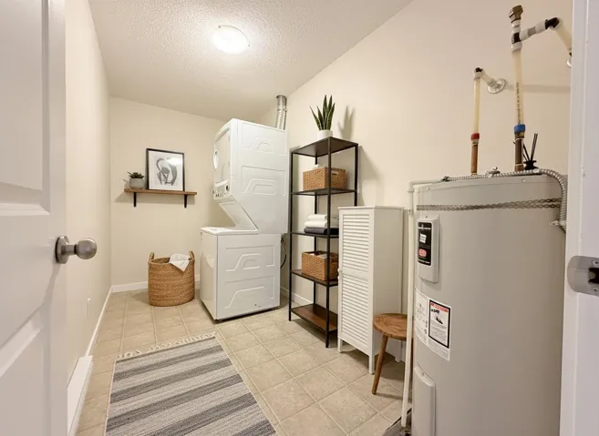 Laundry room featuring electric water heater, stacked washer and clothes dryer, a textured ceiling, and light tile patterned flooring
