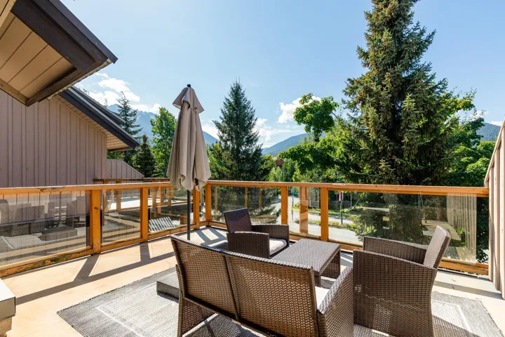 Outdoor deck with wood railings and glass panels, featuring a neutral-toned exterior wall, and a backdrop of mature trees and mountains