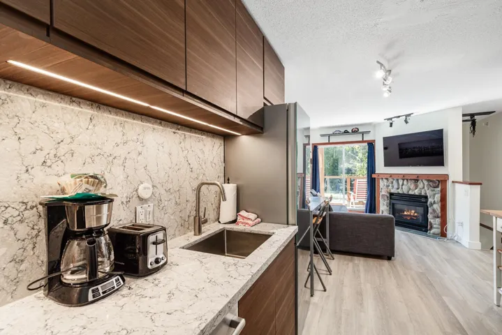 Integrated kitchen and living space featuring wood-finish cabinetry, a stone-finish kitchen backsplash, a stainless steel sink, wood-finish flooring, and a stone-clad fireplace