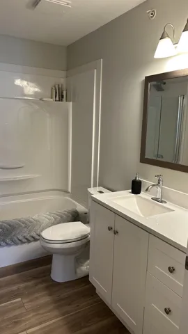 Bathroom featuring a white vanity with integrated basin, light-toned stone-finish countertop, wood-finish flooring, and a tub-shower combination with a surround enclosure