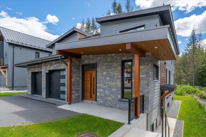 Contemporary exterior featuring a stone and siding facade, covered entry with recessed lighting, integrated two-car garage, wood-finish columns, and a prominent wood-finish entry door with vertical glass inserts