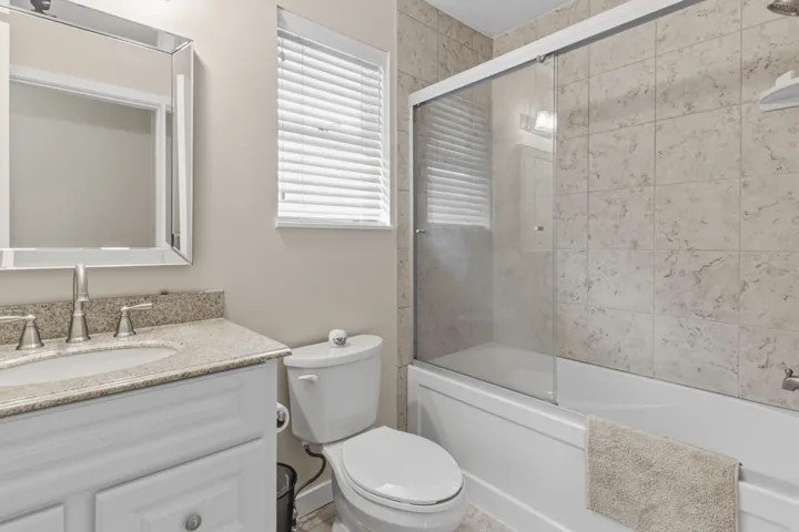 Bathroom featuring a stone-look tiled tub surround, a built-in white bathtub with sliding glass doors, a vanity with a stone-look countertop and integrated sink, and a framed mirror