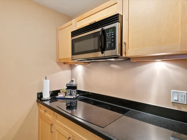 Kitchen with stainless steel microwave, dark countertops, black electric cooktop, and light brown cabinetry