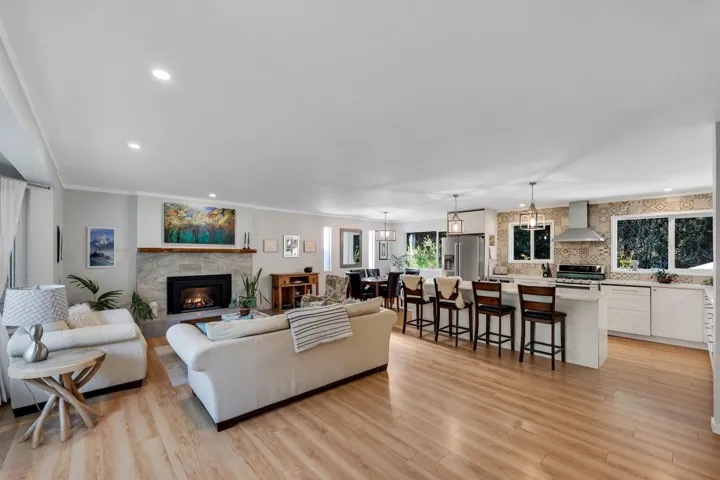 Open-concept living space featuring wood-finish flooring throughout, a fireplace with a stone surround and wood mantel, recessed lighting, and a kitchen with an island, pendant lighting, and a decorative tile backsplash