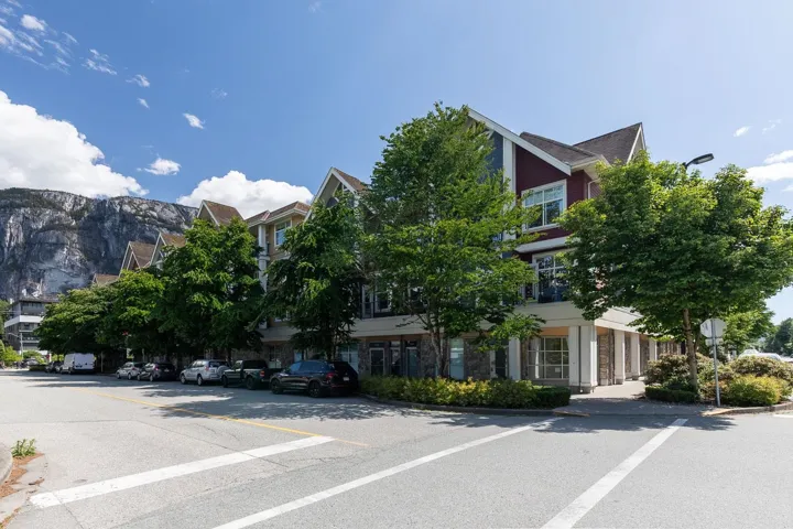 Multi-story residential building featuring gabled rooflines, red and beige siding, stone accents, and mature trees lining the street