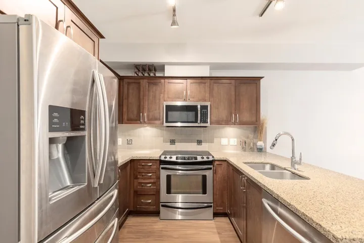 U-shaped kitchen featuring stainless steel appliances, wood-finish cabinetry, speckled countertops, a double basin sink, and a tile backsplash