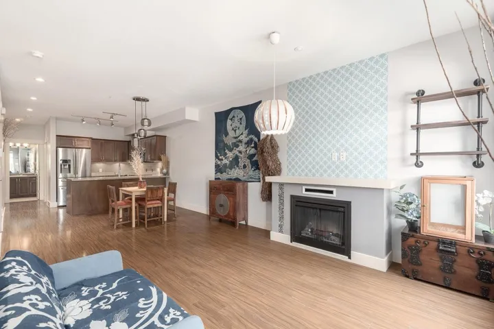 Open-concept living space featuring wood-finish flooring, a fireplace with a light-colored mantel, and a kitchen with dark cabinetry, stainless steel appliances, and a central island