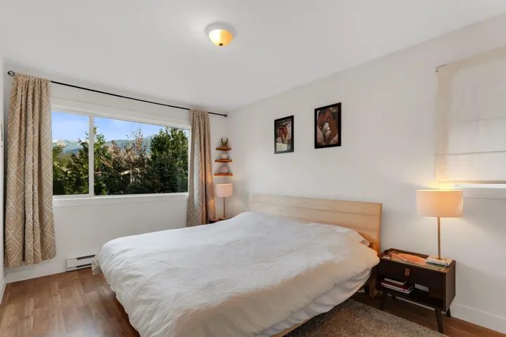 Bright bedroom featuring a large window with mountain and tree scenery, wood-finish flooring, white walls, and a ceiling-mounted light fixture
