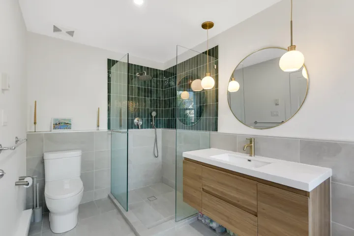 Contemporary bathroom featuring a floating wood-finish vanity with a white countertop and undermount sink, a large circular mirror with brass-finish frame, and an enclosed shower with dark green vertically-stacked subway tiles and a rainfall showerhead