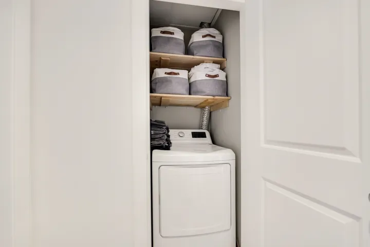 Dedicated laundry closet featuring a white dryer with a digital display, two wood-finish storage shelves, and a white paneled door