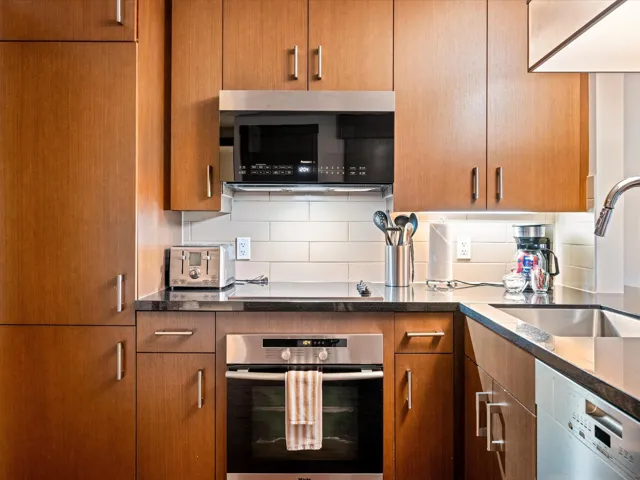Kitchen featuring stainless steel appliances, tasteful backsplash, brown cabinetry, and dark stone countertops