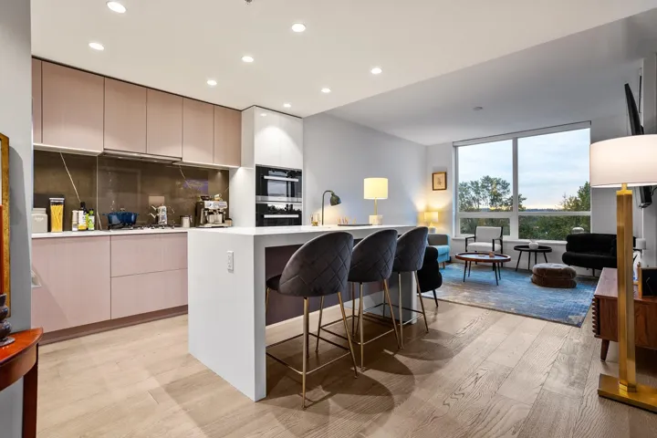 Open-concept living space with wood-finish flooring, a kitchen island with seating, integrated wall oven, and large windows