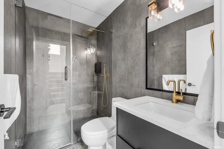 Modern bathroom featuring a glass-enclosed shower with gold-tone fixtures, a built-in toilet, a vanity with an integrated sink and gold-tone faucet, a rectangular mirror, and a contemporary light fixture