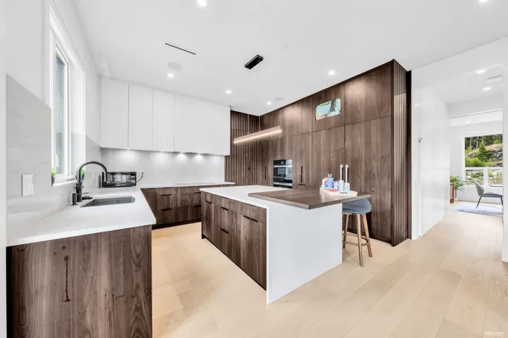 Gourmet kitchen featuring a central island with integrated seating, built-in wall oven and microwave, dark wood-finish cabinetry, white upper cabinetry, and light wood-finish flooring