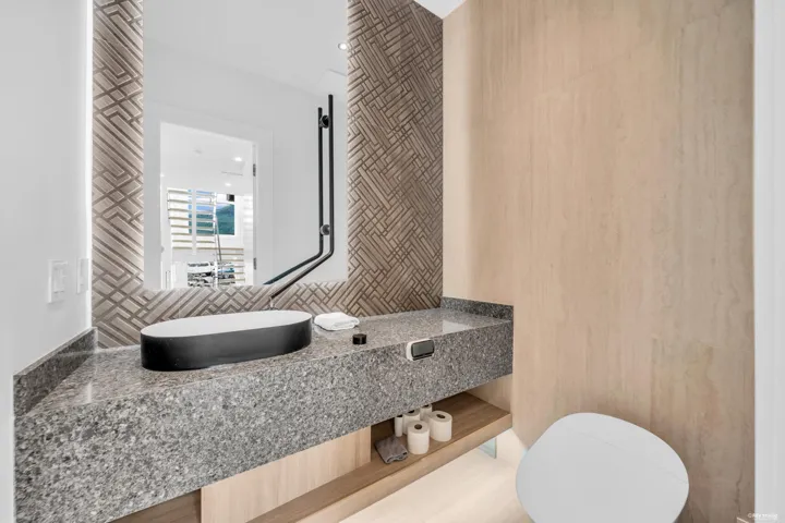Contemporary bathroom featuring a vessel sink on a speckled gray countertop, a mirror with geometric-patterned trim, and light wood-finish cabinetry