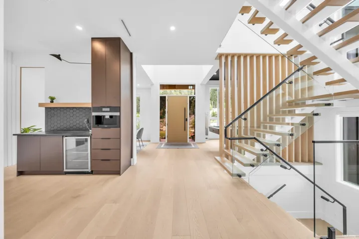 Open-concept entry area featuring light wood-finish flooring, a contemporary floating staircase with glass railings, an integrated beverage station with dark cabinetry, an entryway with a large wooden door, and vertical wood slat accents