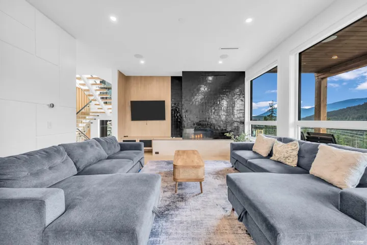 Spacious living area featuring large windows showcasing mountain scenery, a contemporary fireplace with a dark tiled surround, light wood-finish flooring, and a modern open staircase with glass railings