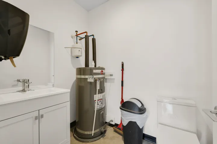 Utility space featuring a water heater, white vanity with integrated sink, chrome faucet, wall-mounted mirror, and a white toilet