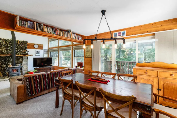 Open-concept living and dining area featuring a stone fireplace with a wood-burning stove, wall-to-wall windows, a built-in shelf system, wood-paneled walls, and a glass sliding door providing outdoor access