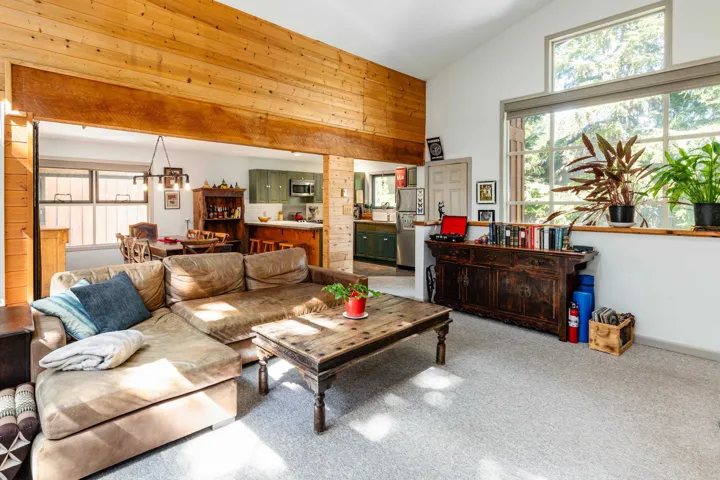 Spacious living area featuring a vaulted ceiling with a wood-clad accent wall, large picture windows, light-toned carpeting, and a visible dining area