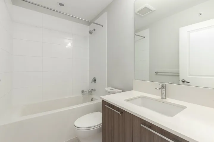 Contemporary bathroom featuring a white bathtub, white tiled shower surround, a white integrated sink, a light-toned countertop, and wood-finish vanity cabinetry with brushed nickel hardware