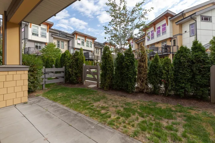 Rear exterior featuring a concrete patio, landscaped yard with grass and evergreen shrubs, and two wooden gates