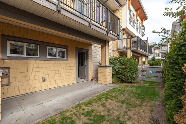 Ground-level entry featuring light yellow shingle siding, a concrete walkway, two transom windows with white frames, and a privacy fence bordering a grassy area