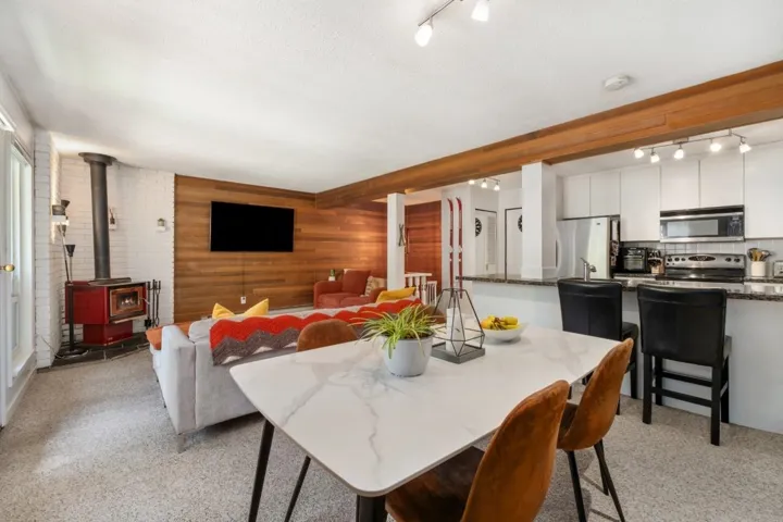 Open-concept living area featuring a white marble-look dining table, wood-beam accents, a red freestanding stove, a wood-paneled accent wall, and a kitchen with stainless steel appliances and dark countertops