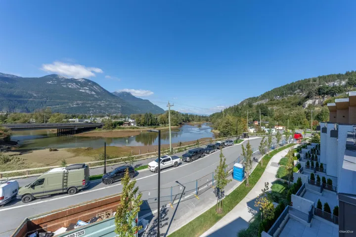 Riverfront property featuring a paved road, pedestrian walkway, mature mountains, and a modern building exterior