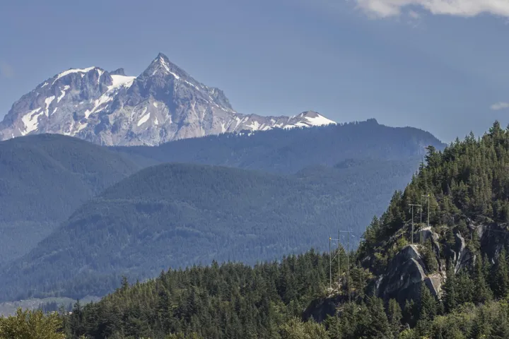 Snow-capped mountains rise above a dense forest landscape under a clear blue sky