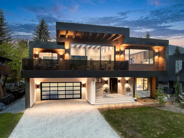 Modern architectural exterior featuring a prominent two-story design with extensive glazing, a multi-paneled garage door, a stone veneer facade, a second-story balcony with metal railing, and wood-finish paneling accents