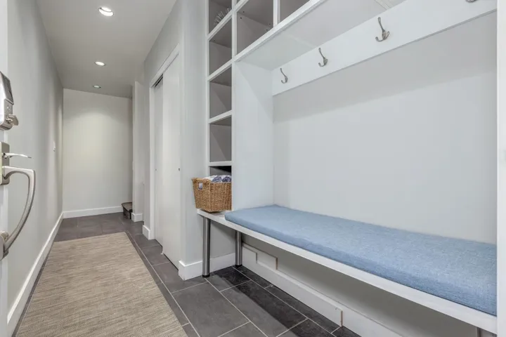 Entryway featuring built-in storage with open shelving, a built-in bench with a light blue cushion, wall-mounted coat hooks, recessed lighting, and dark grey tile flooring
