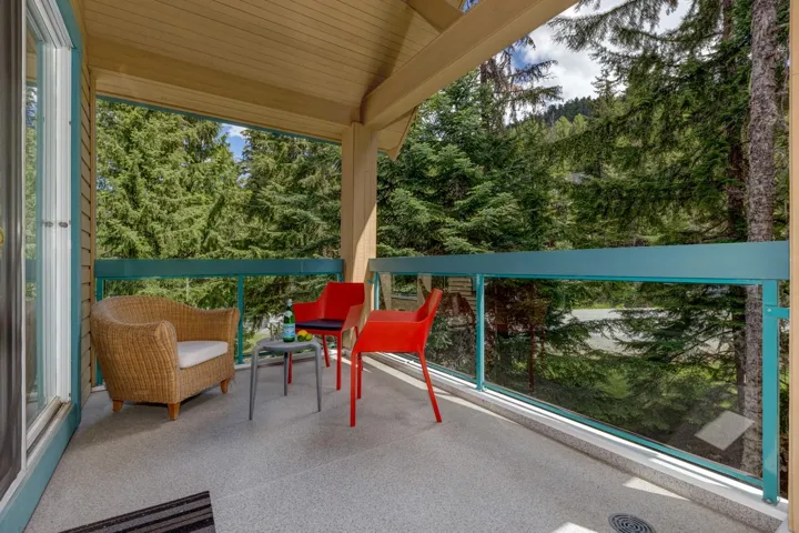 Outdoor balcony featuring glass panel railing, composite decking, a wood-paneled ceiling, and surrounding mature trees