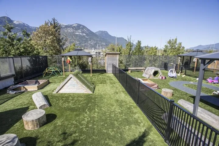 Fenced outdoor space with artificial turf, multiple play structures, a sandbox, and a covered gazebo