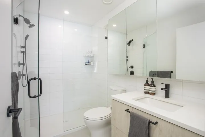 Walk-in shower with a glass enclosure and matte black fixtures, white subway tile surround, wood-finish vanity with a white countertop and integrated sink, and an expansive mirrored medicine cabinet