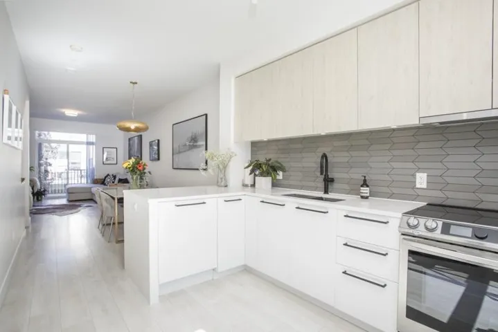 Modern kitchen featuring white cabinetry with integrated hardware, quartz countertops, a gray subway tile backsplash, stainless steel appliances including a slide-in range and built-in oven, and a waterfall-edge island with seating
