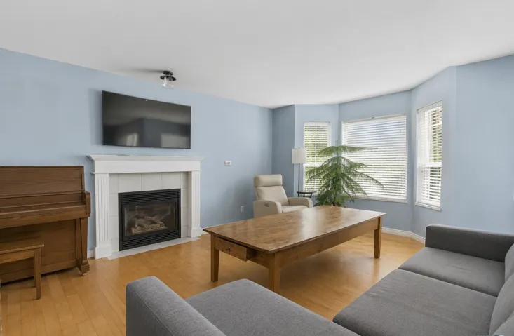 Spacious living area featuring light wood-finish flooring, a white mantel fireplace with tile surround, and multiple windows with horizontal blinds