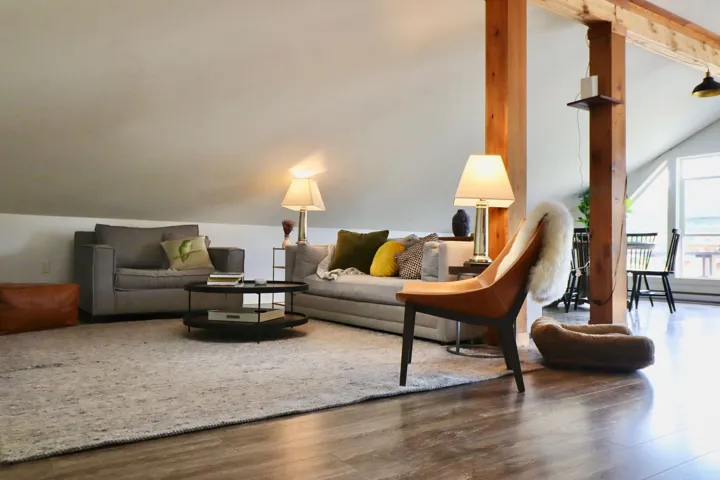 Spacious living area featuring wood-finish flooring, exposed wood beams, sloped ceilings, a large area rug, and ample natural light from multiple windows