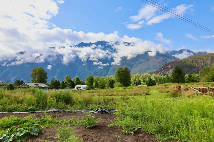 Expansive agricultural land featuring a greenhouse structure, a barn-style building, and distant mountain ranges with cloud cover