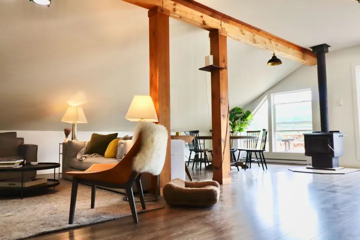 Open-concept living area featuring exposed wood beams, wood-finish flooring, a freestanding wood-burning stove, and vaulted ceilings with a prominent triangular window
