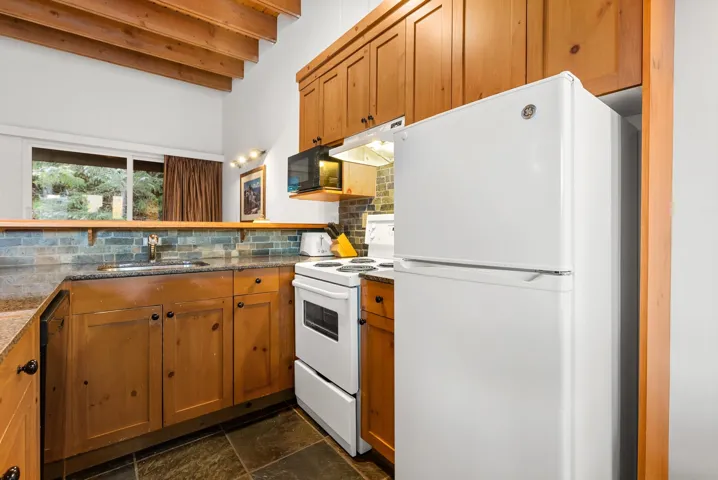 Kitchen with white appliances, backsplash, beam ceiling, brown cabinetry, and under cabinet range hood