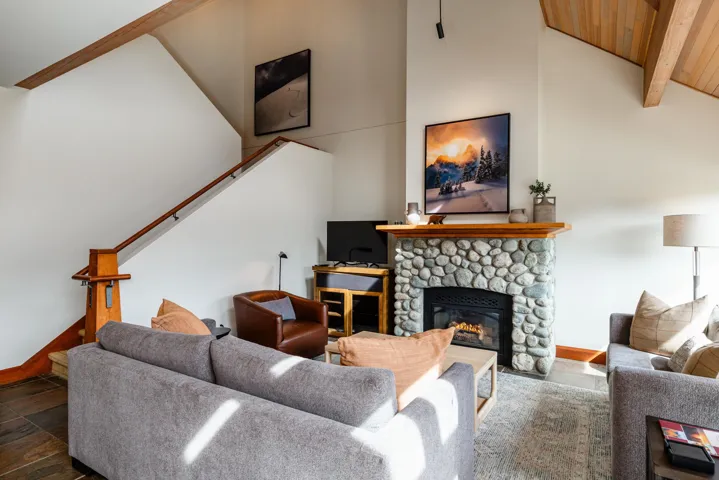 Living area featuring a stone fireplace with a wood mantle, vaulted ceilings with exposed wood beams, a wood-finish staircase, and dark tile flooring