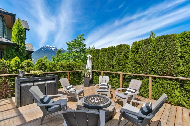 Expansive wood-finish deck featuring a built-in fire pit, mountain backdrop, and tall privacy hedges