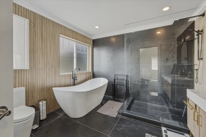 Modern bath featuring a freestanding soaking tub with floor-mounted faucet, a spacious glass-enclosed shower with rain head, dark large-format floor tiles, wood-paneled wall accents, and recessed lighting