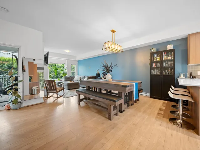 Open-concept living space featuring light wood-finish flooring, a contemporary gold-tone chandelier, a dedicated dining area, a defined living area with large windows, and a kitchen island with built-in bar seating